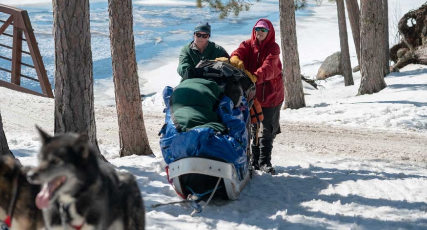Two mushers push a packed sled across snow. There are sled dogs in the foreground. 